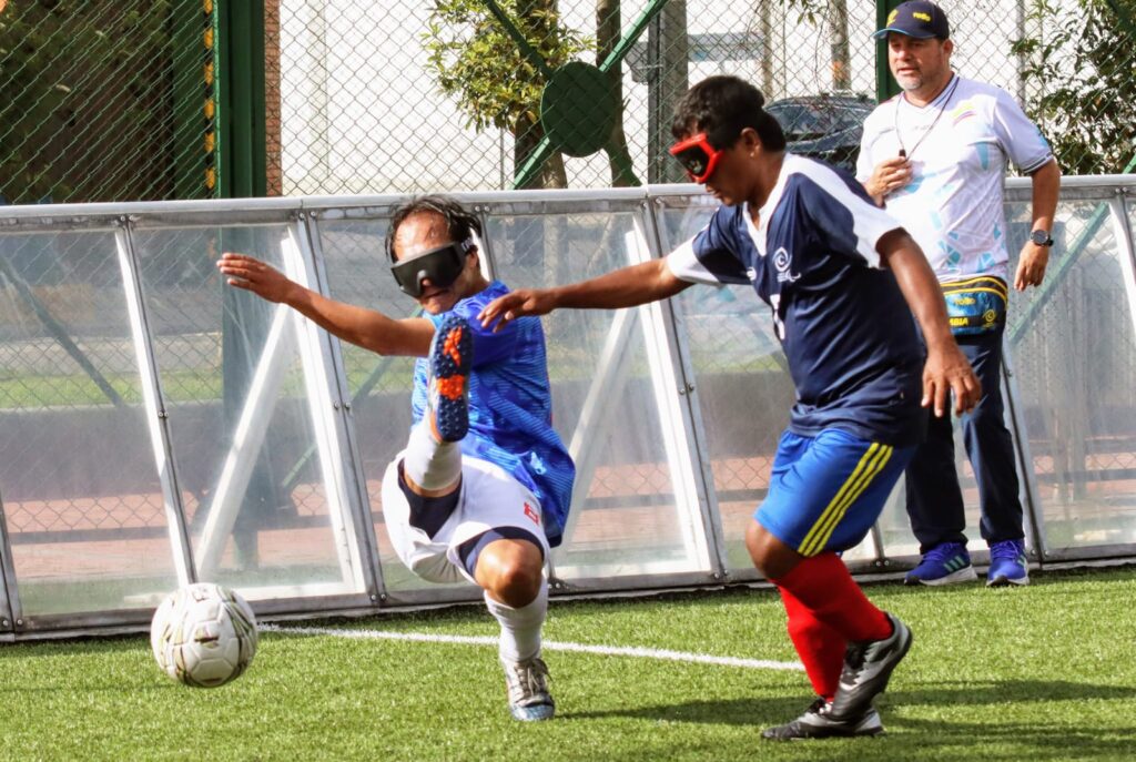 Juador de Fútbol para ciegos pateando la pelota, portando gafas especiales del deporte. Lleva puesta una pantaloneta blanca y una camiseta azul. 