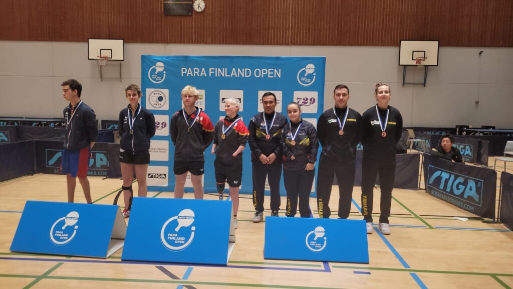 Ocho deportistas participantes del Abierto de Para Tenis de mesa de Finlandia,  ubicados en fila, con los brazos detras de la espalda, portando una medalla. De fondo, una pancarta azul con los logos de los patrocinadores del evento